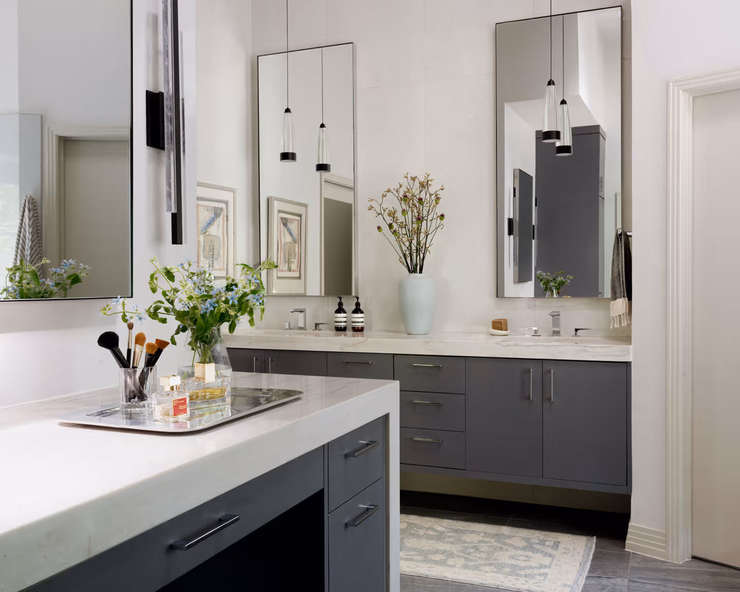 Modern kitchen with gray cabinets, marble countertops, island with bar stools, built-in ovens, and gray tile flooring.