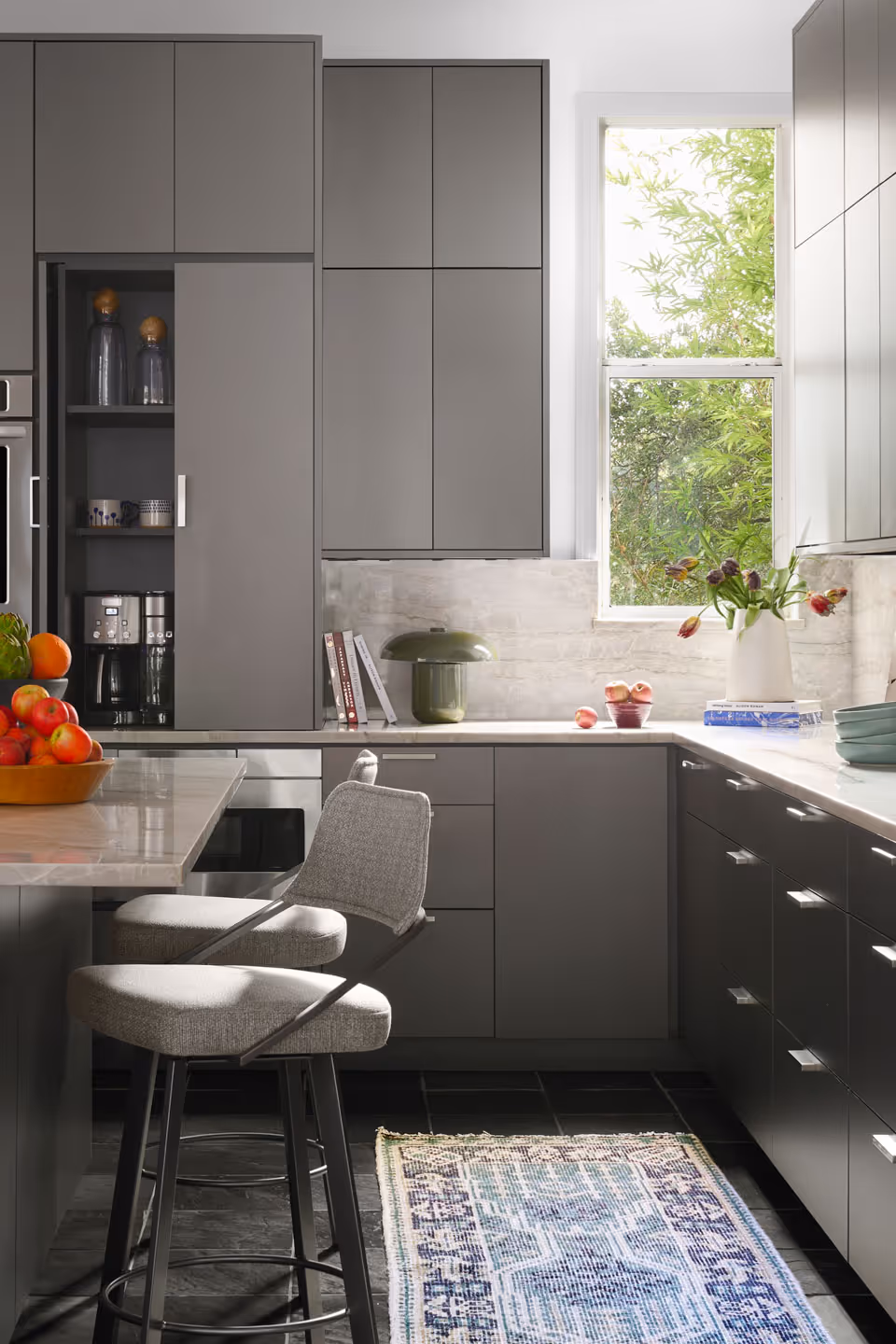 Modern kitchen with gray cabinets, marble countertop, two gray bar stools, and a window with green plants outside.