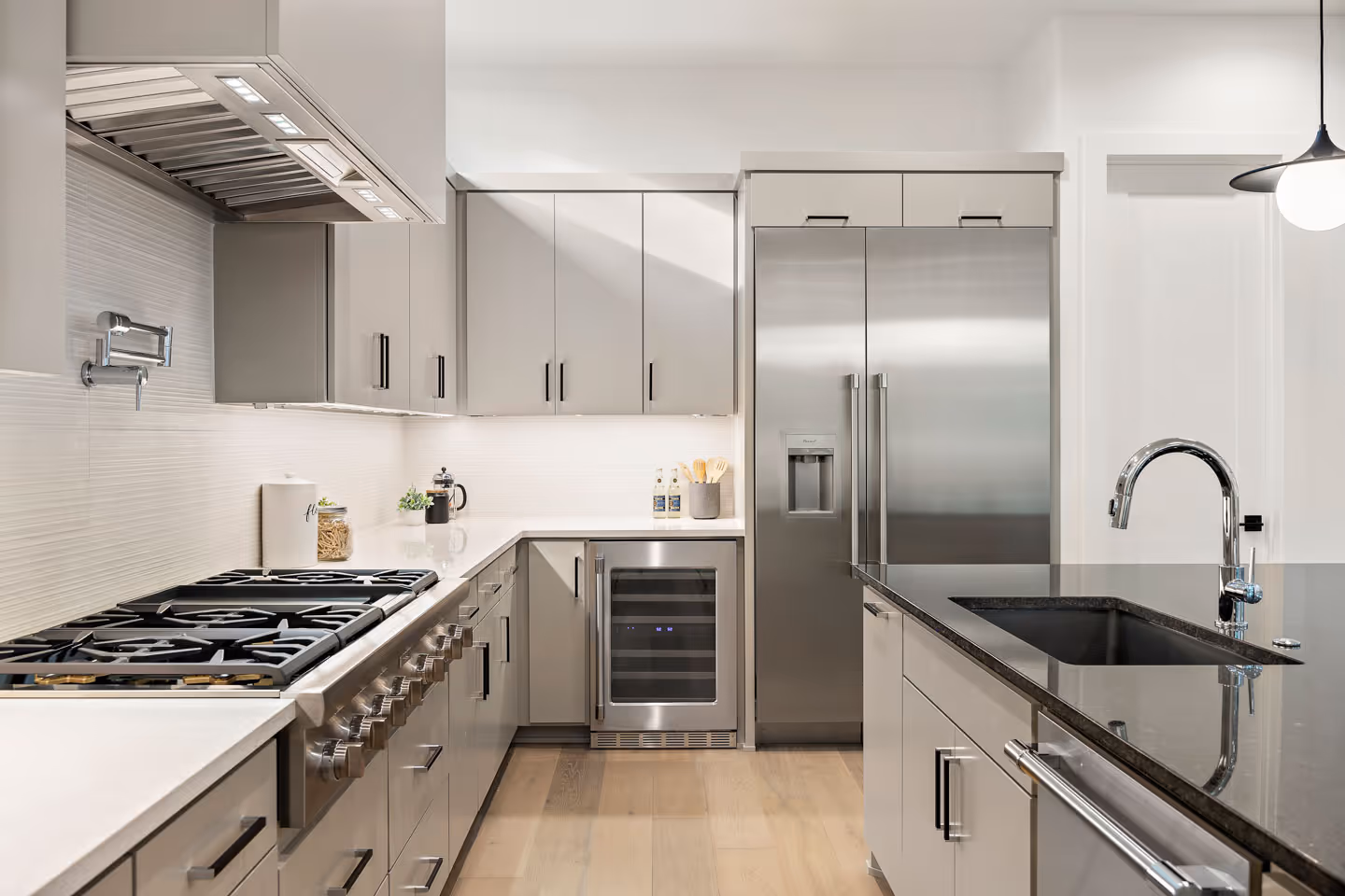 Modern kitchen with stainless steel appliances including a gas stove, wine cooler, and double-door refrigerator, and a black countertop island with a sink and faucet.