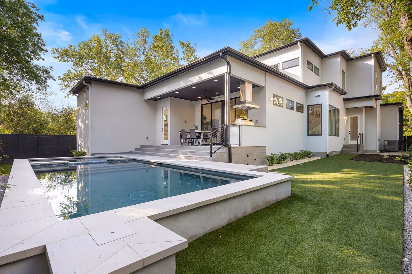 Modern two-story white house with a covered patio, outdoor dining area, built-in grill, and a rectangular swimming pool surrounded by green lawn.
