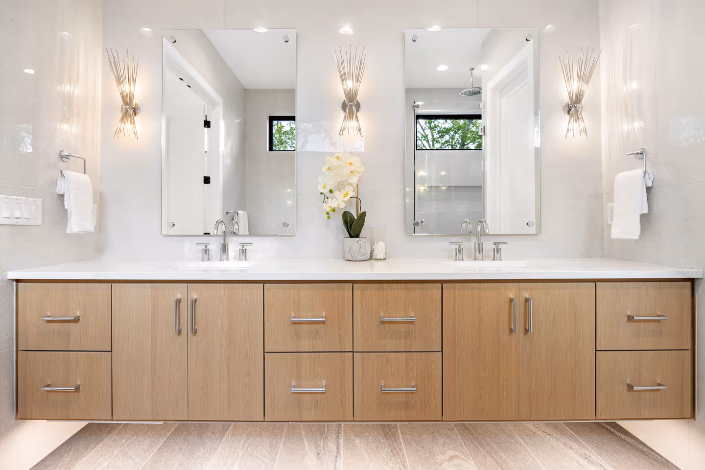 Modern double vanity bathroom with light wood cabinets, two mirrors, stainless faucets, white countertop, wall sconces, and white orchid centerpiece.