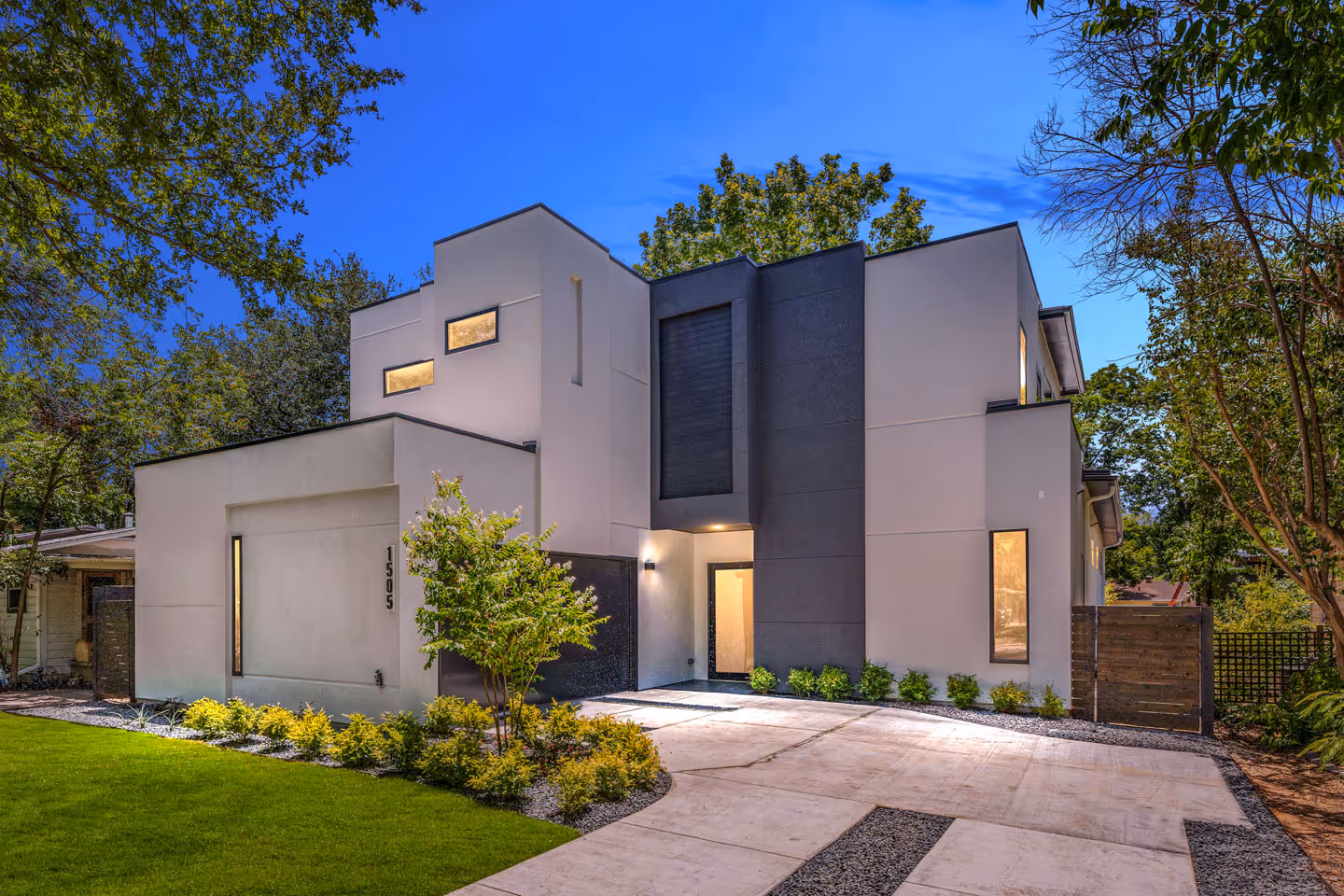 Modern two-story house with a flat roof, gray and dark exterior walls, illuminated windows, and a concrete driveway surrounded by greenery at dusk.