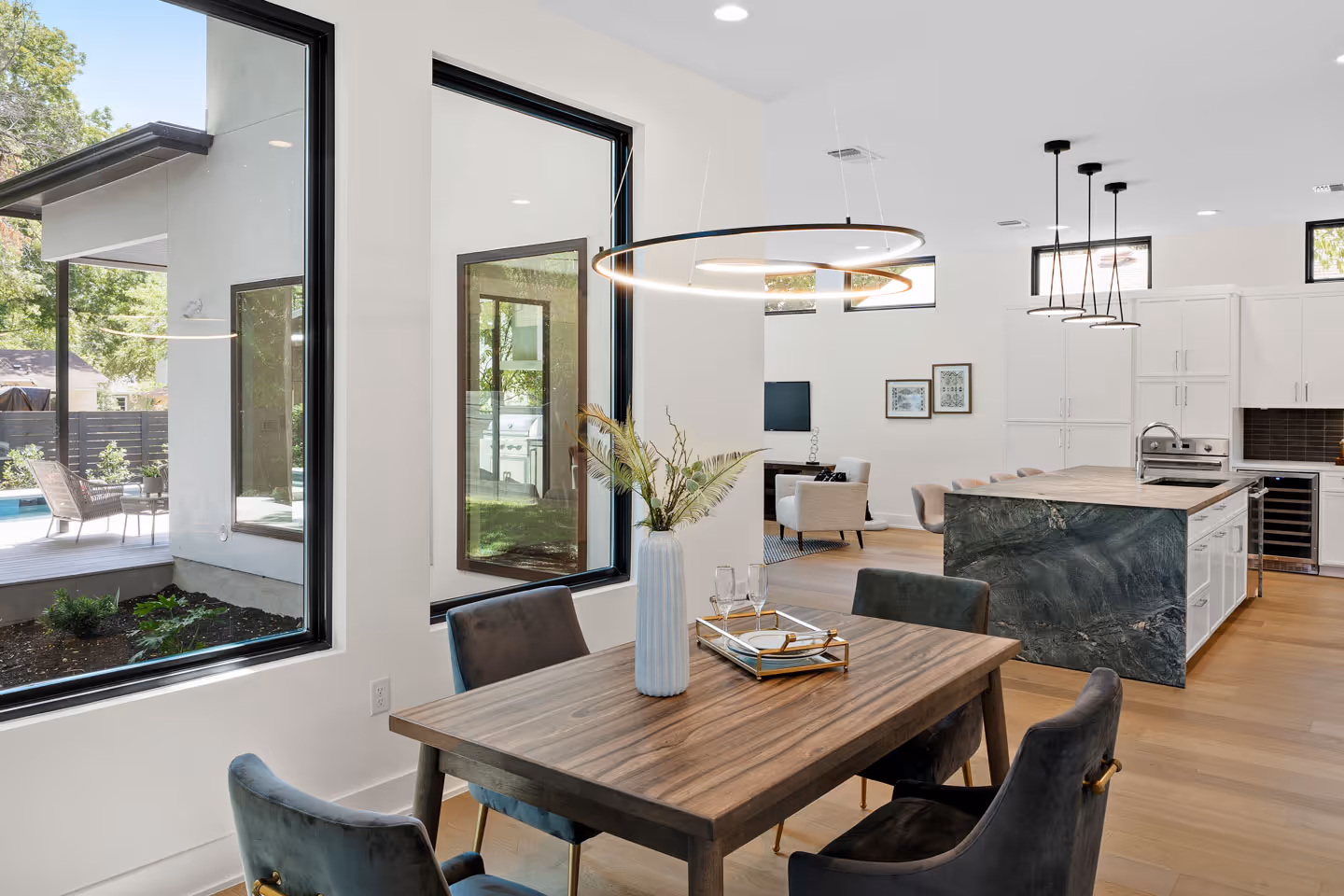 Modern open-concept kitchen and dining area with wooden table, velvet chairs, a decorative vase, large windows, and an island with pendant lights.