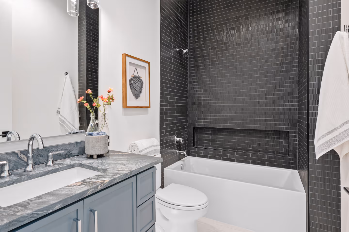 Modern bathroom with a white bathtub surrounded by black tiled walls, gray marble countertop with sink, blue cabinetry, and decorative pink flowers in a vase.
