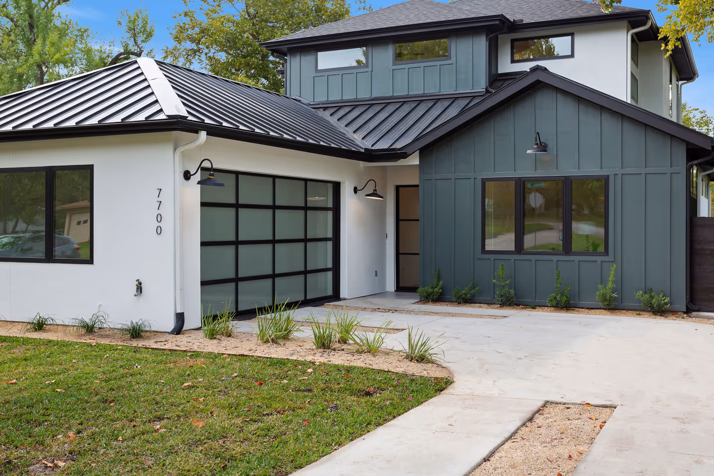 Modern two-story house with dark metal roofs, white and gray exterior walls, glass garage door, and landscaped yard.