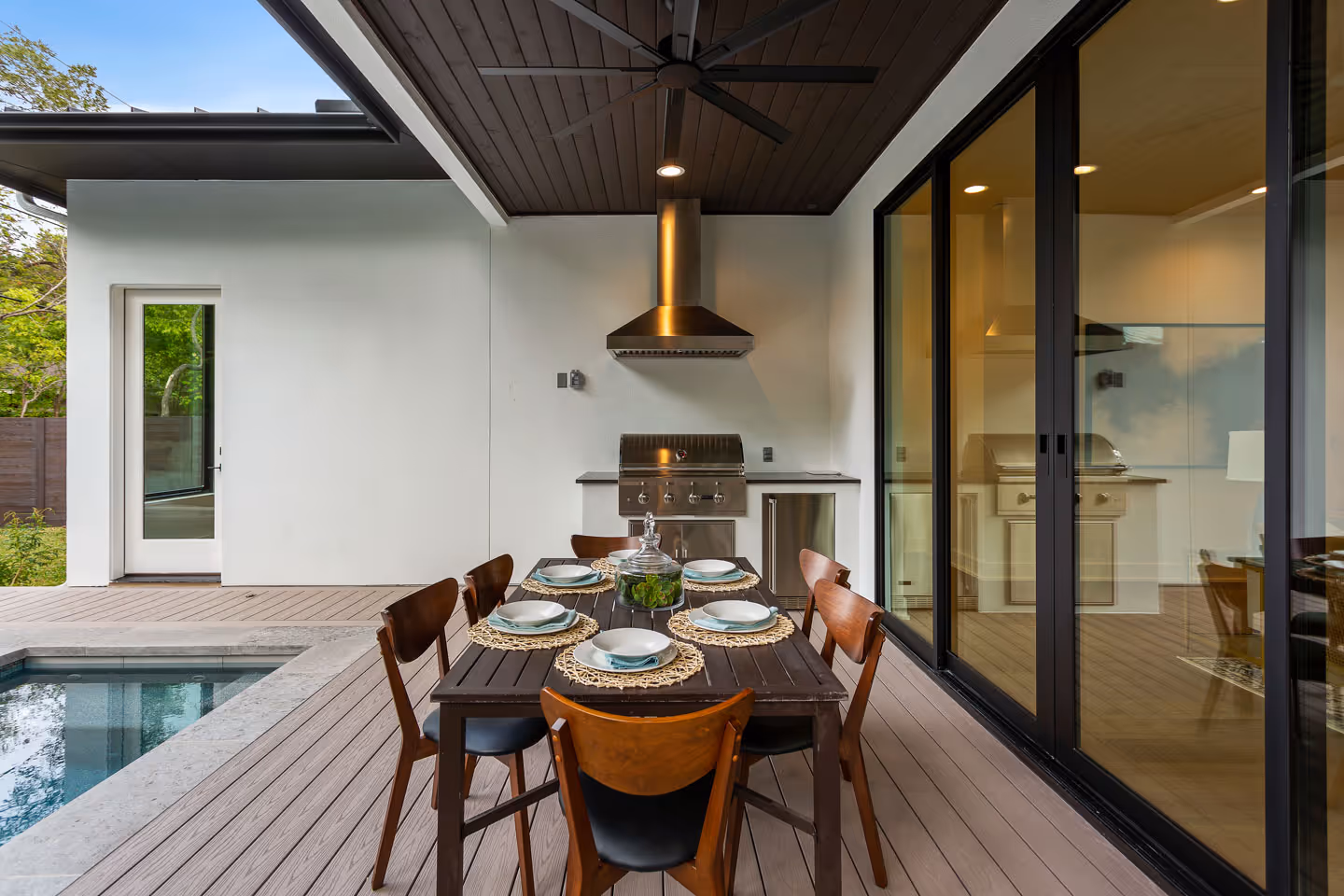 Modern outdoor dining area with a wooden table set for six, a stainless steel grill under a ceiling fan, and a pool beside the deck.