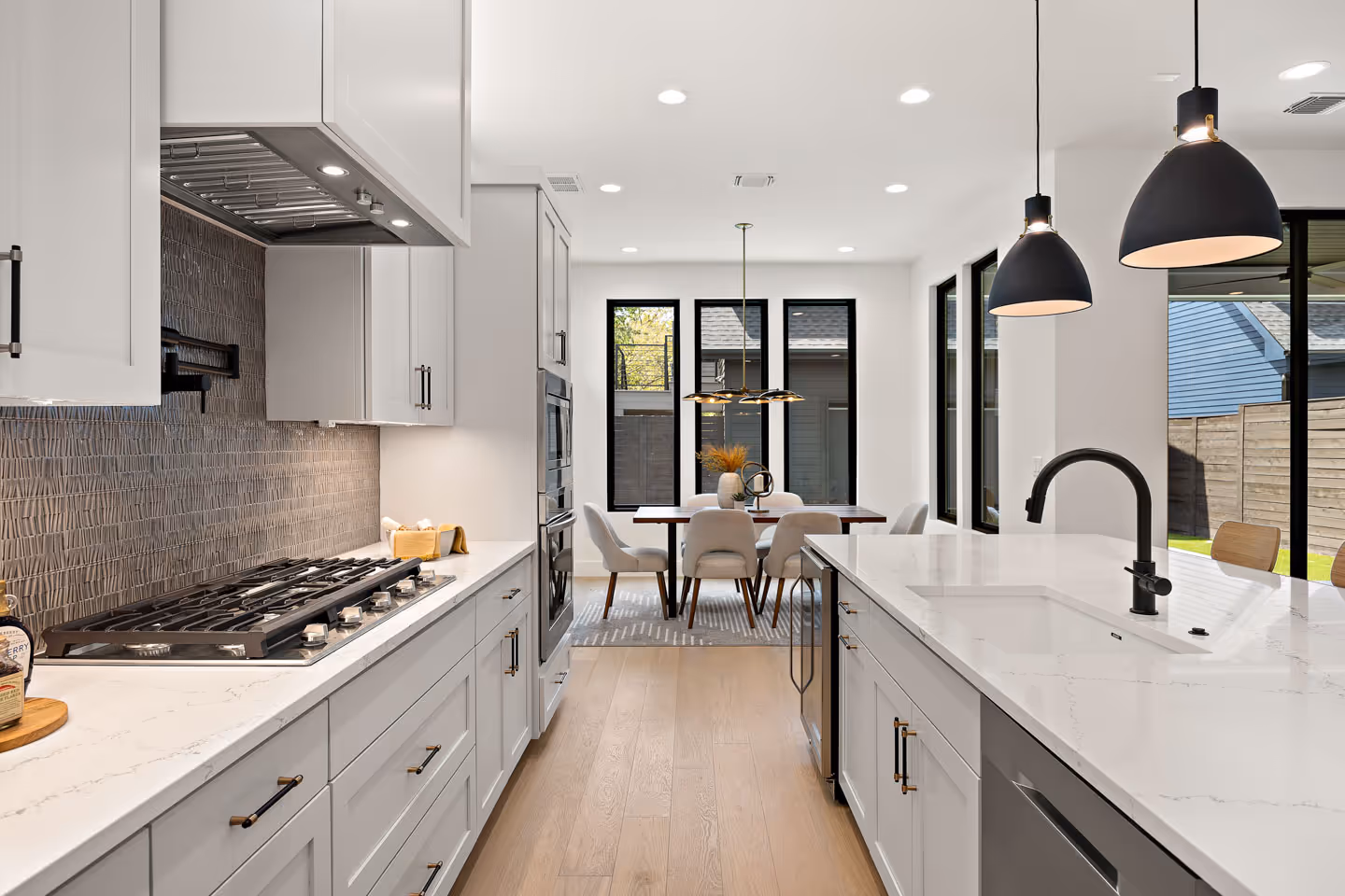 Modern kitchen with white cabinetry, marble countertops, black fixtures, and a dining area with large windows and beige chairs.