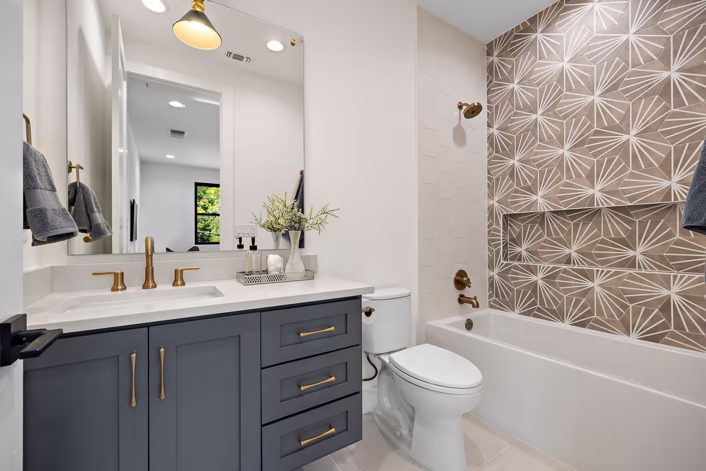 Modern bathroom with gray vanity, brass fixtures, white countertop, toilet, and bathtub with geometric patterned tile wall.