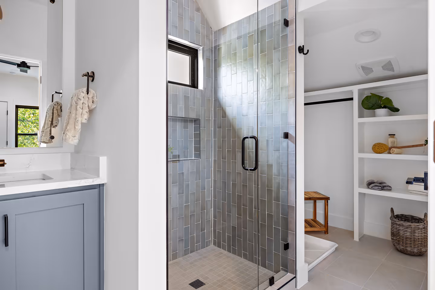 Modern bathroom with a glass shower enclosure featuring vertical gray tiles, a blue vanity with white marble countertop, and open shelving with decor and towels.