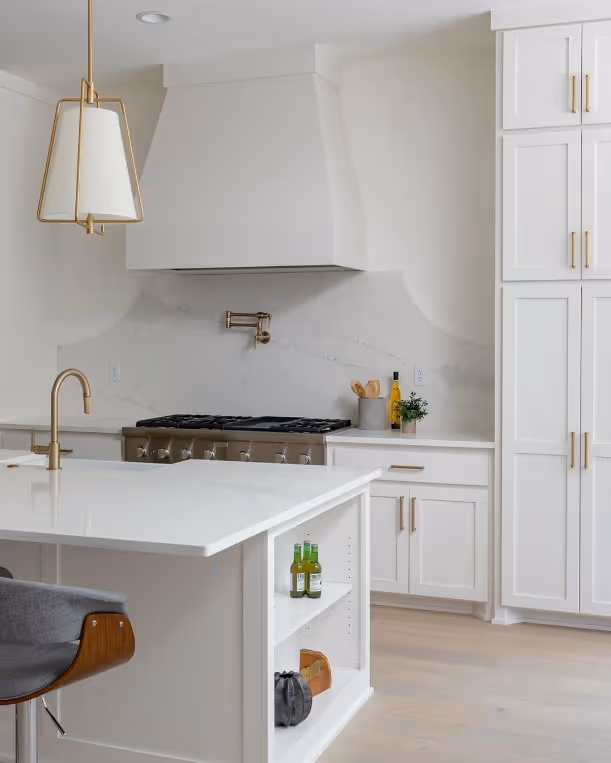 Modern white kitchen with a large island, brass fixtures, built-in shelving, and a gas stove with a white range hood.