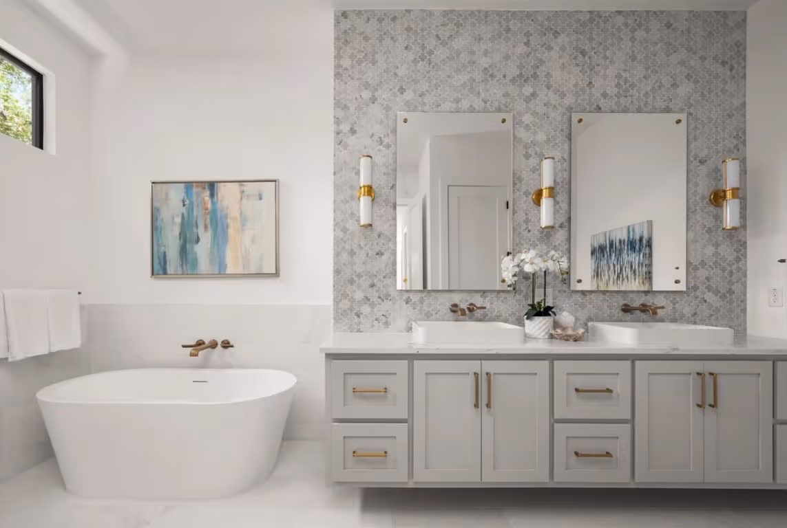Modern bathroom with freestanding white bathtub, gray double vanity with vessel sinks, two mirrors, gold fixtures, and mosaic tiled wall.
