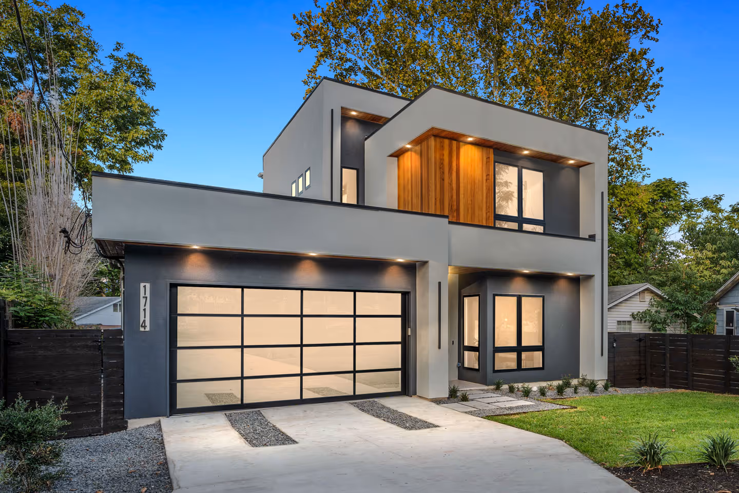Modern two-story house with gray and white exterior, wooden accents, large glass garage door, and illuminated windows at dusk.
