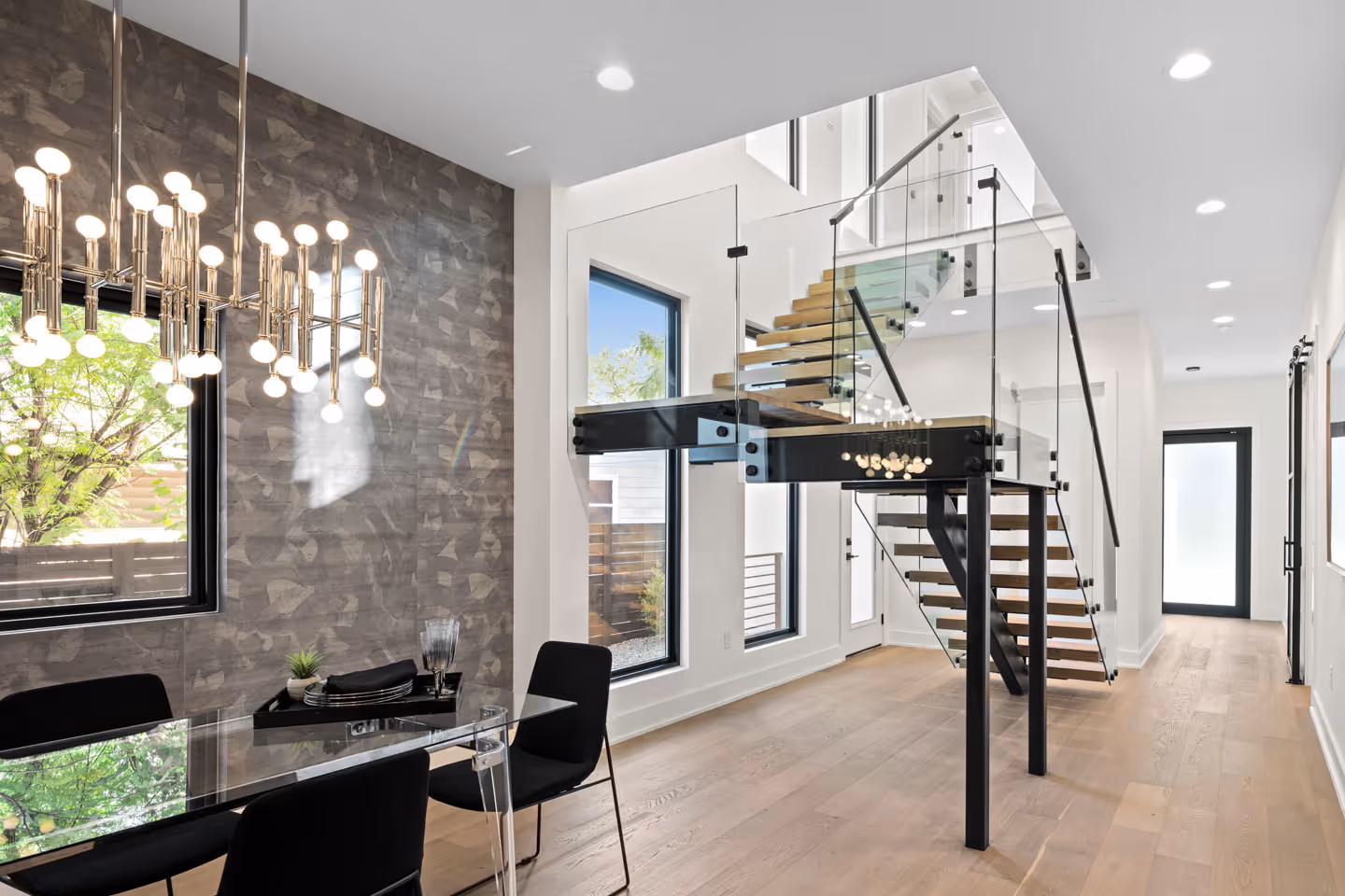 Modern interior with glass dining table, black chairs, a chandelier, and a floating staircase with glass railings.
