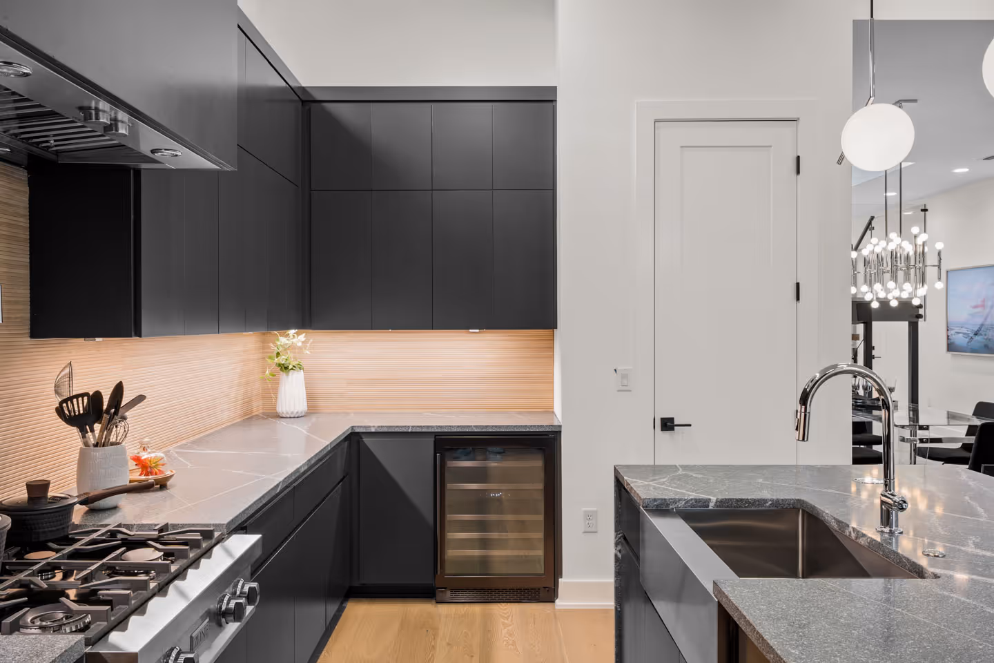 Modern kitchen with black cabinets, gray stone countertops, built-in wine cooler, stainless steel sink, and wooden floor.