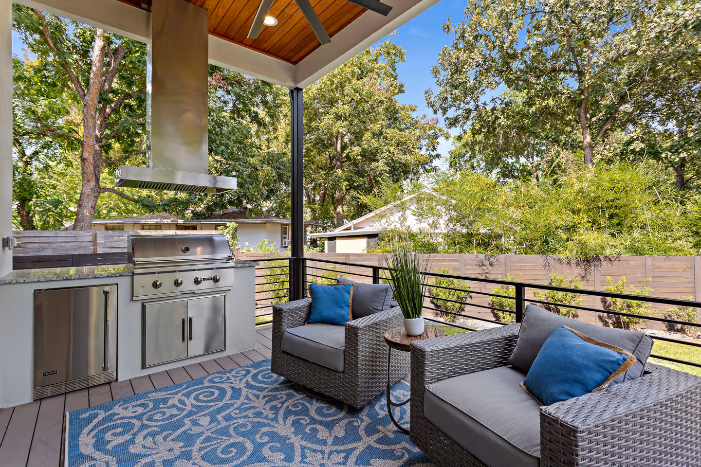 Outdoor patio with built-in stainless steel grill, two gray wicker armchairs with blue pillows, a small round wooden table with a potted plant, and a blue patterned rug.
