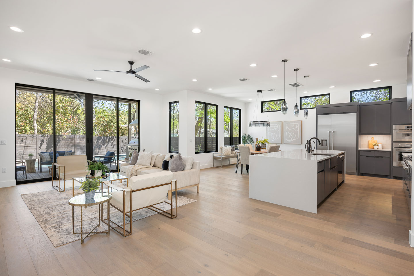 Modern open-plan living space with light wood floors, white walls, beige seating area, a large kitchen island with black cabinetry, dining table, and large windows overlooking a fenced backyard with trees.