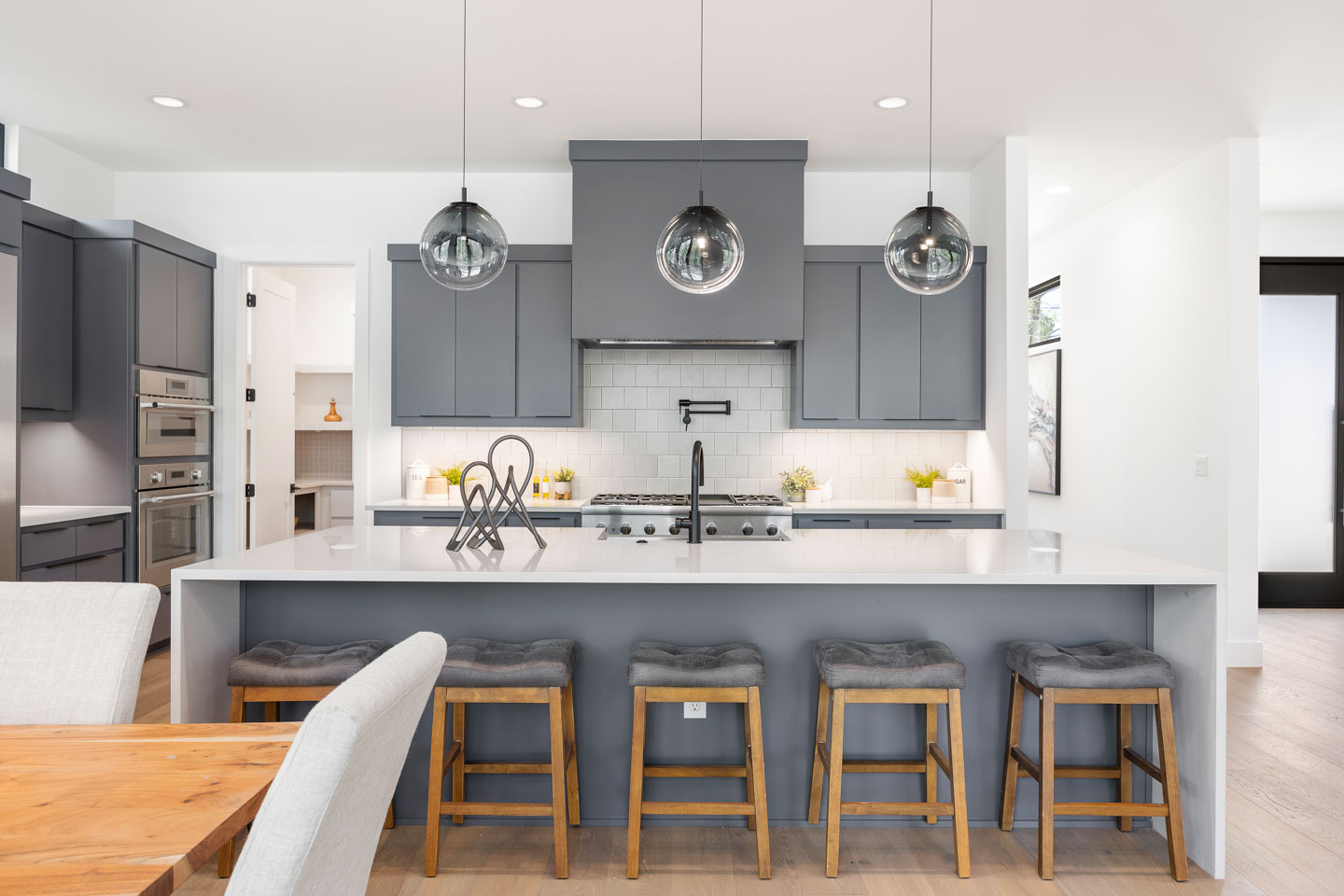 Modern kitchen with gray cabinets, white countertops, five wooden stools with gray cushions at the island, and three glass pendant lights.