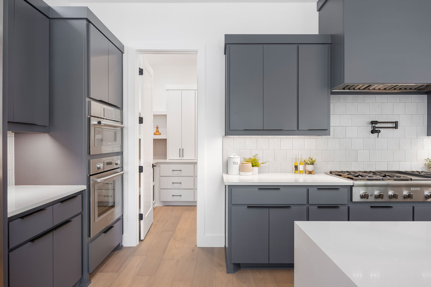 Modern kitchen with gray cabinets, stainless steel oven and stove, white subway tile backsplash, and light wood flooring.