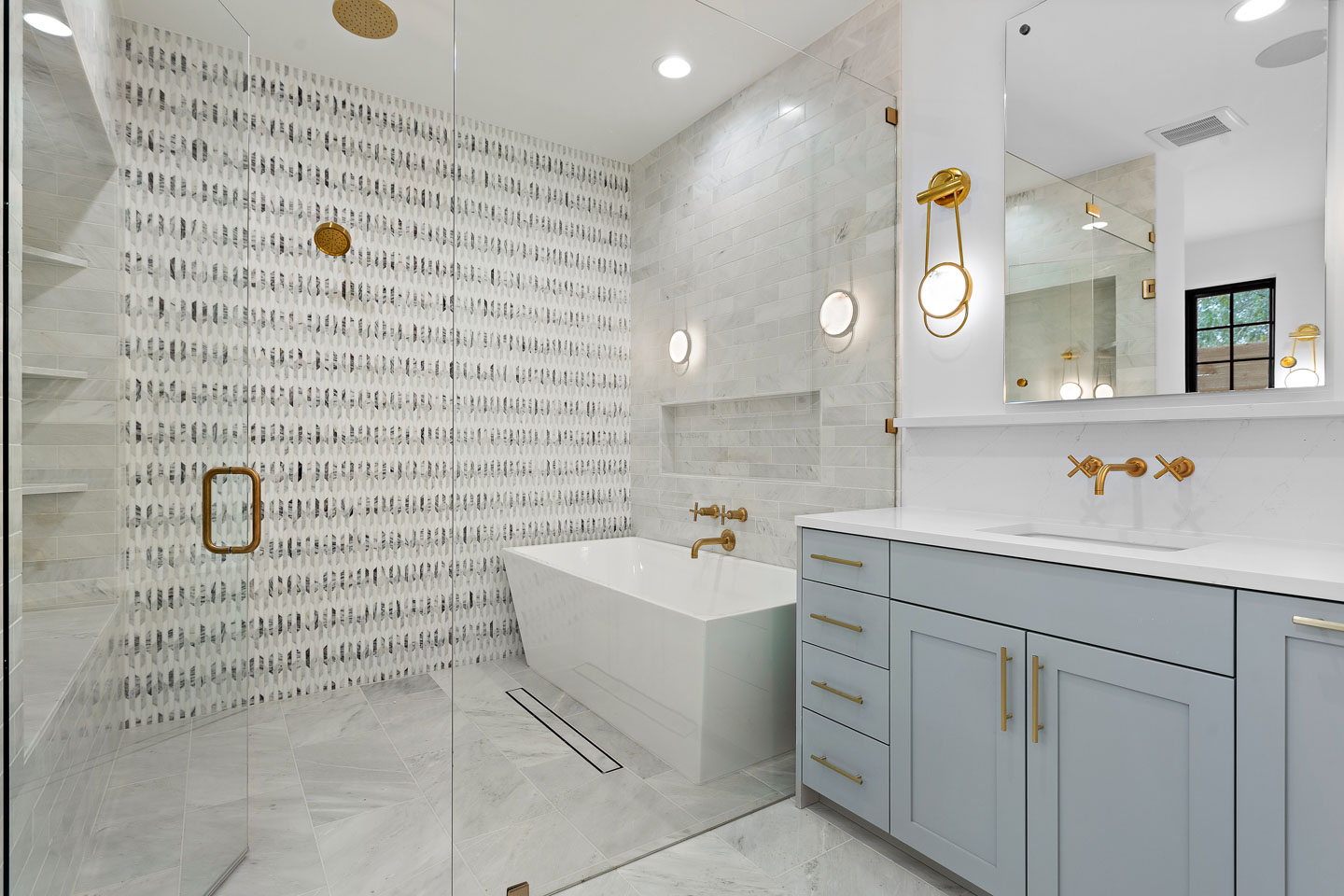 Modern bathroom with gray tile floor, a glass-enclosed shower, freestanding white bathtub, light blue vanity with gold fixtures, and patterned accent wall.
