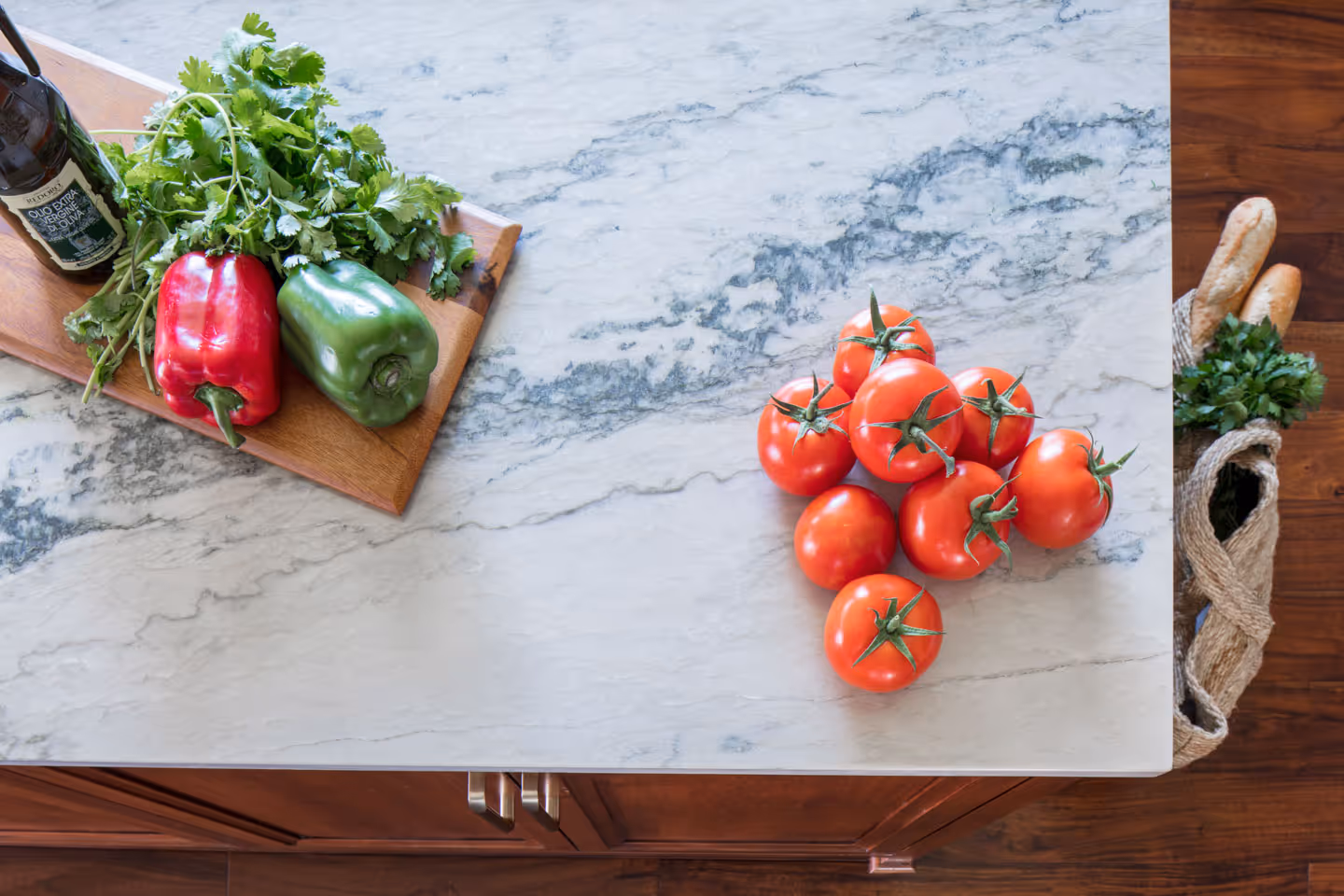 Wooden kitchen countertop with a cutting board holding green and red bell peppers and parsley, tomatoes on the marble surface, and a woven grocery bag on a rug with baguettes and parsley inside.