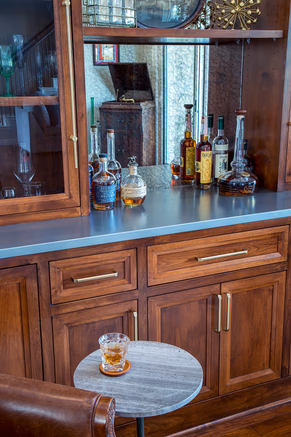 Glass cabinet with several whiskey bottles on a countertop and a glass of whiskey on a small round table in front of a brown leather chair.