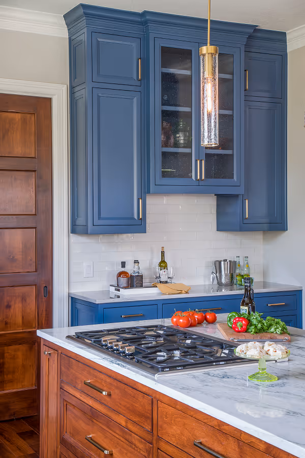 Modern kitchen with blue cabinets, marble countertops, wooden island with cooktop, and hanging pendant lights.