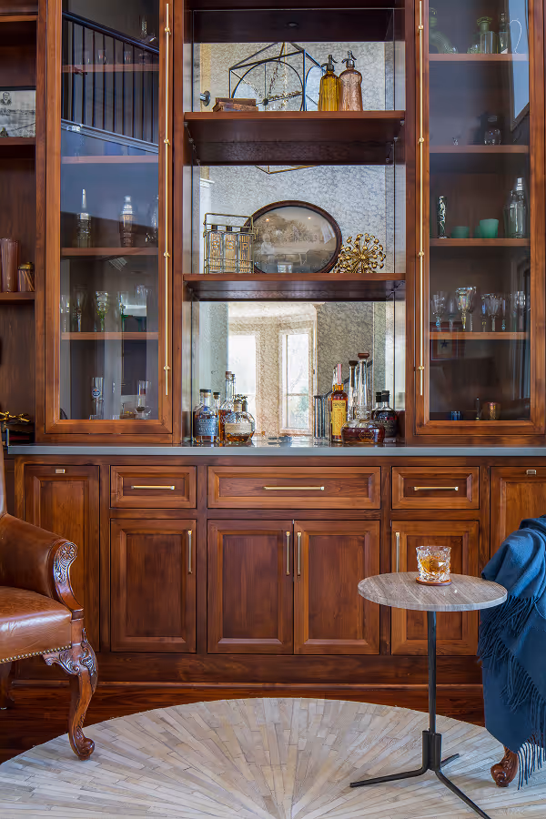 A wooden cabinet with glass doors displaying various bottles and decorative items, next to a leather armchair and a small round table with a glass of whiskey.