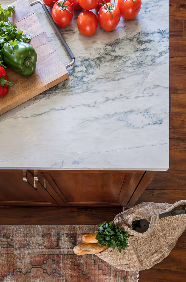Wooden kitchen countertop with a cutting board holding green and red bell peppers and parsley, tomatoes on the marble surface, and a woven grocery bag on a rug with baguettes and parsley inside.