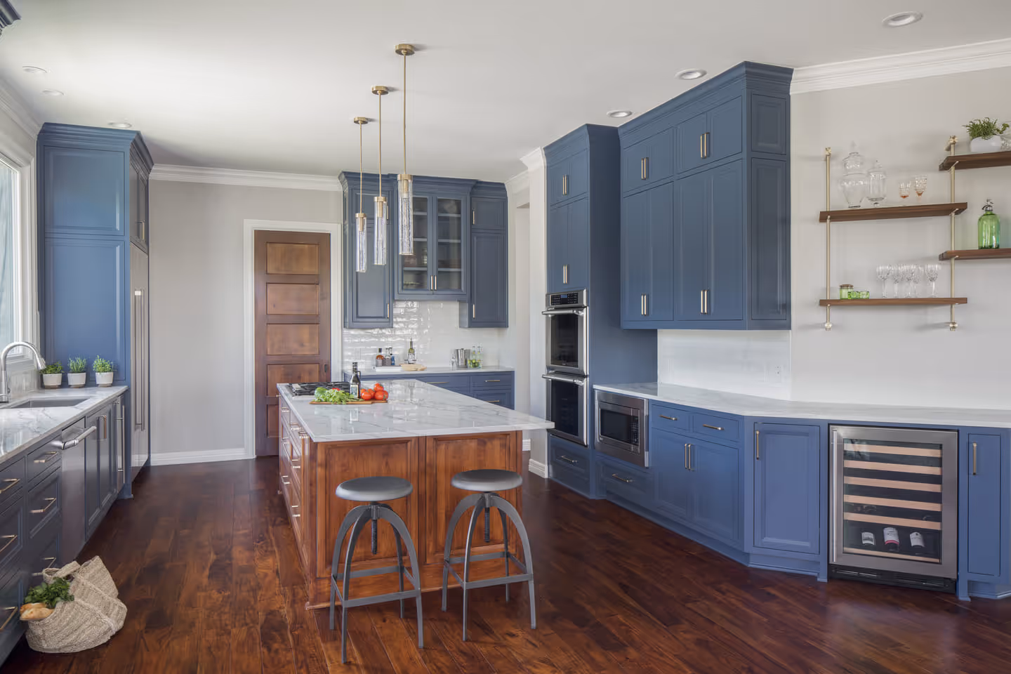 Modern kitchen with blue cabinets, white marble countertops, wooden island with two black stools, pendant lights, and dark hardwood floors.