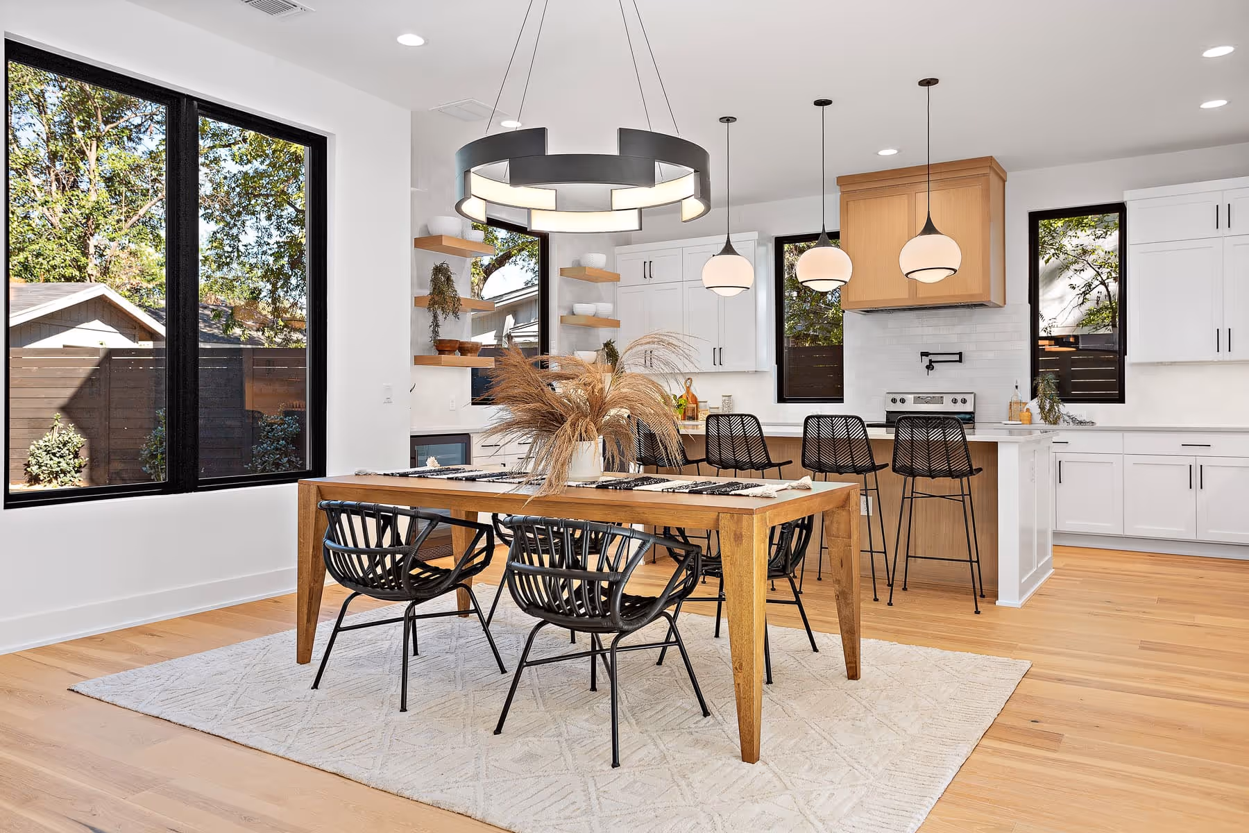 Modern dining area with wooden table, black chairs, large windows, and a kitchen in the background featuring white and wood cabinetry.